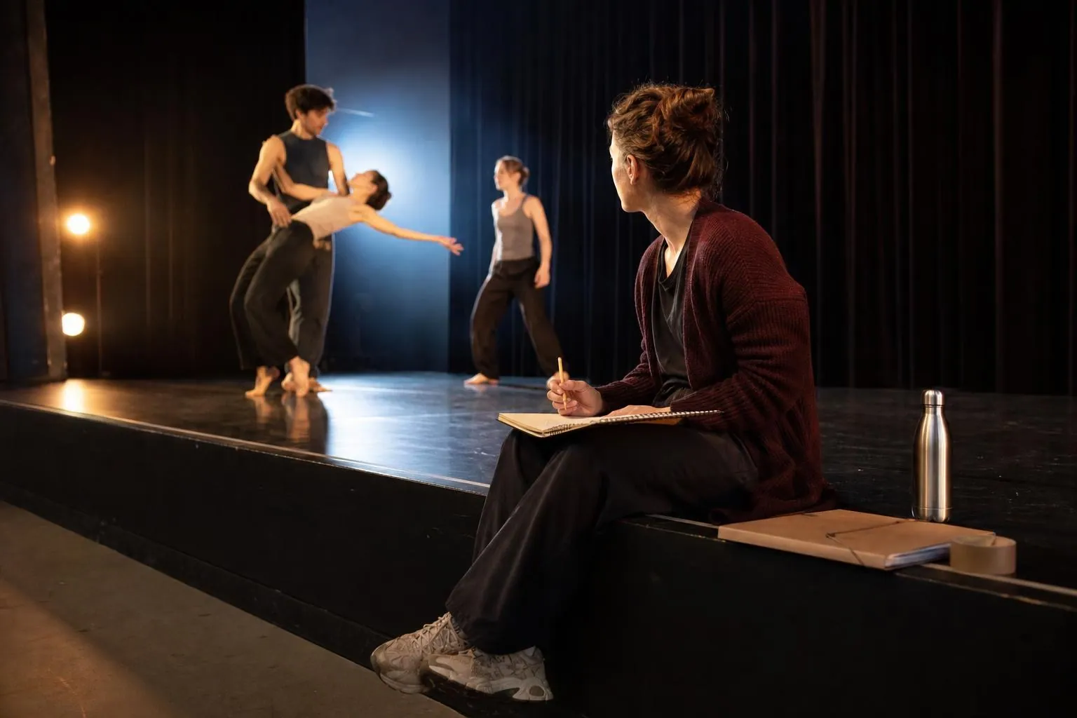 Chargée de production assise au bord du plateau, carnet sur les genoux, pendant une répétition de danse en arrière plan