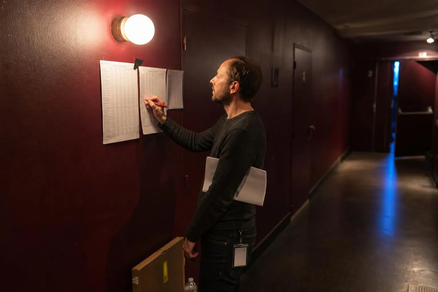 Homme corrigeant au stylo rouge une colonne de chiffres sur un tirage papier punaisé au mur d’un couloir de loges